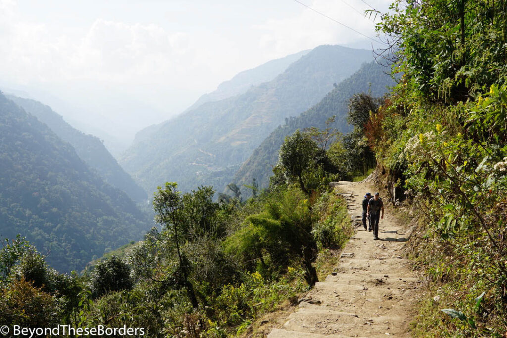 Two hikers walking along the dirt and rock trail with the valley off to their side and green hills behind them.