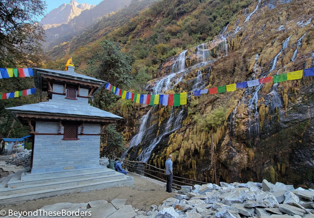 Small temple with prayer flags stung from it outward.  Gentle waterfalls cascading down the hill behind it.  Tall mountains farther off in the distance.