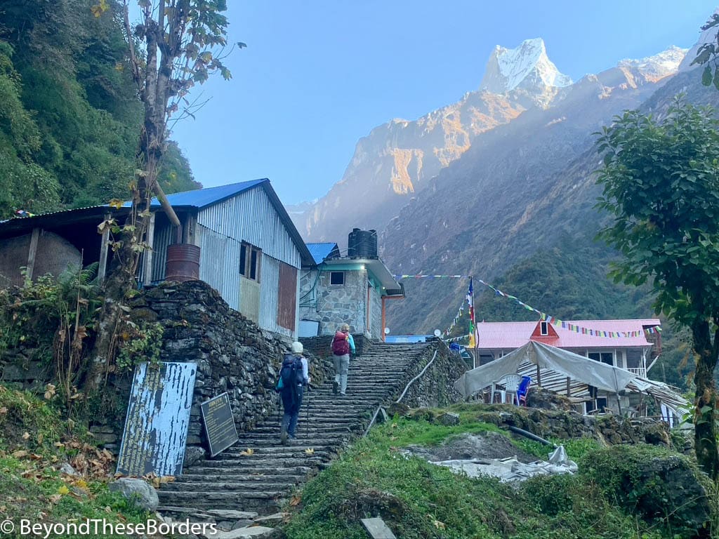 Photo of us hiking up stairs next to simple buildings into a village.  The white peak of Fishtail (mountain) rising hight above in the distance.
