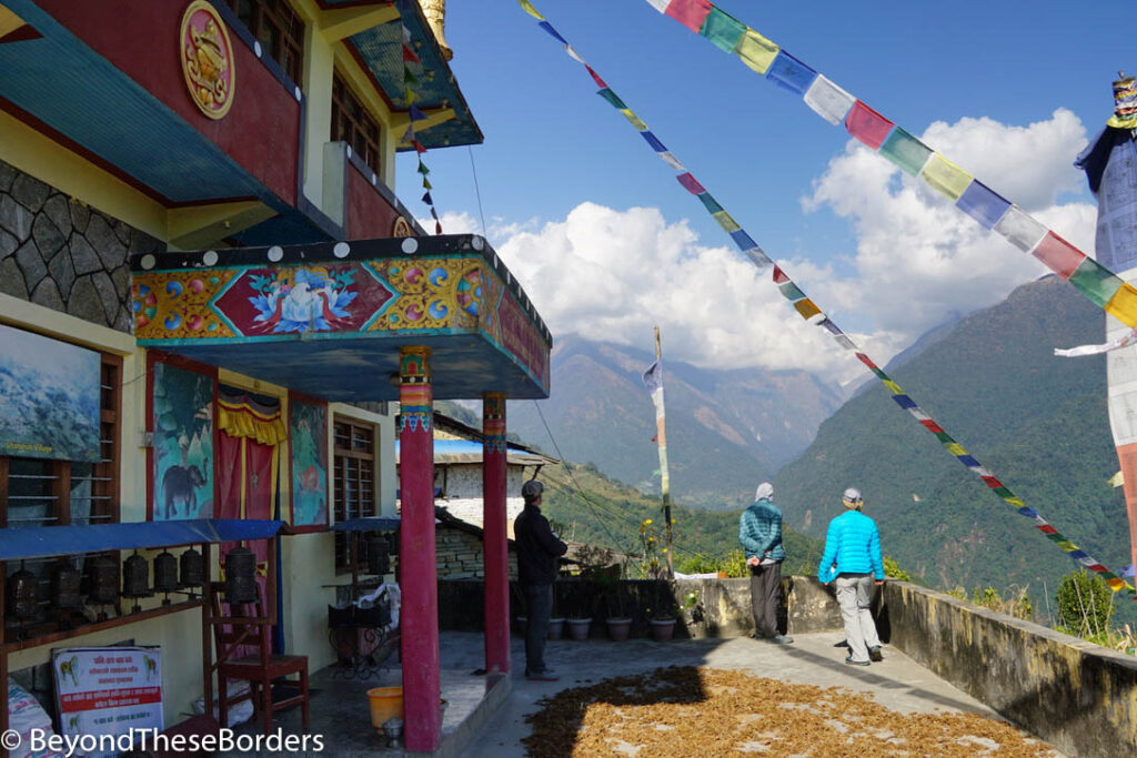Red, blue, and gold temple with prayer flags draped out from it.  Millet like crop drying on the concrete in front of the doors.  View of cloud covered moutons far off in the distance.