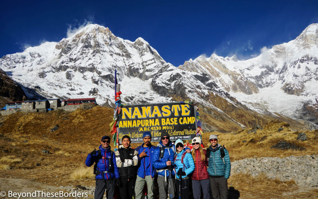 Our group including our guide and 2 porters standing in front of the "Namaste.  Annapurna Base Camp (4130 meters)" sign and clear view of base camp and the Annapurna range behind us in the morning.