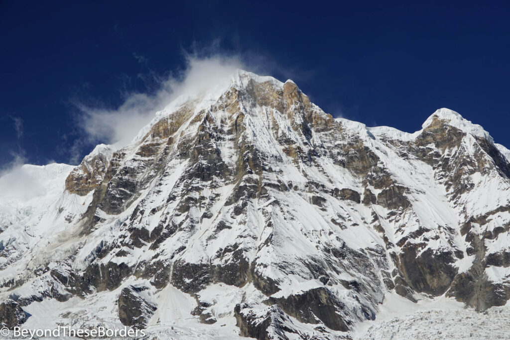 Close up of Annapurna South.  Dark grey and brown jagged rocks near peak with about 50% snow coverage.  Small amount of fog hovering along left side of peak.