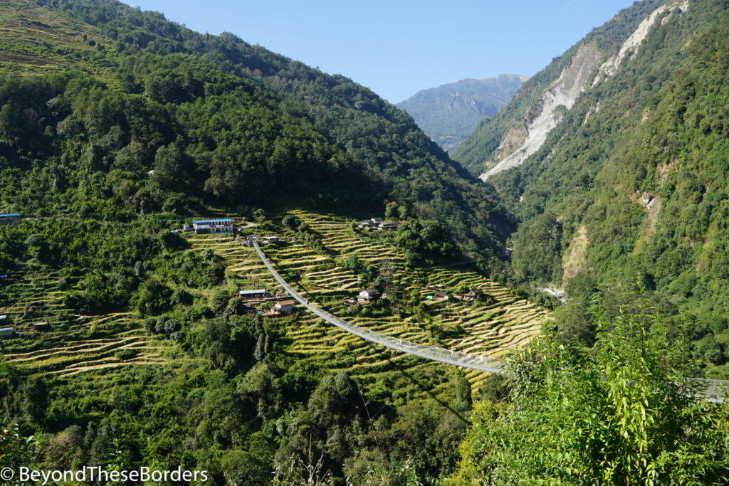 Long steel suspension bridge over deep valley.  Green terraced hill on other side.