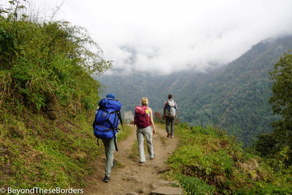 Two hikers and a porter carrying a large bag on the dirt trail. Clouds cover most of the green tree covered hills around the trail.